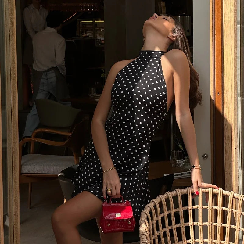 Woman in a polka dot dress holding a red handbag, standing in a room with chairs and a door.