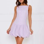 A woman wearing a black sleeveless mini dress with a square neckline and a-line silhouette, paired with a pink handbag and white pearl bracelet.