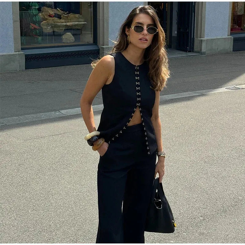 A woman wearing a sleeveless black top with button details and wide-leg pants, both high-waisted. She is accessorized with sunglasses, a watch, and carrying a bag.