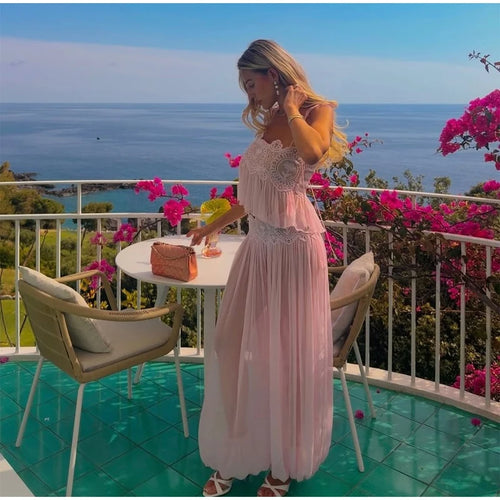 A woman stands on a balcony overlooking the ocean, wearing a light pink sleeveless maxi dress with lace detailing. She is holding a small pink clutch bag next to a white table with two chairs, with bright pink bougainvillea flowers in the background.