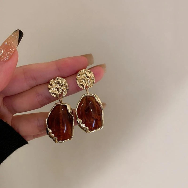 Gold and brown earrings held by a hand with glittery nail polish on a neutral background