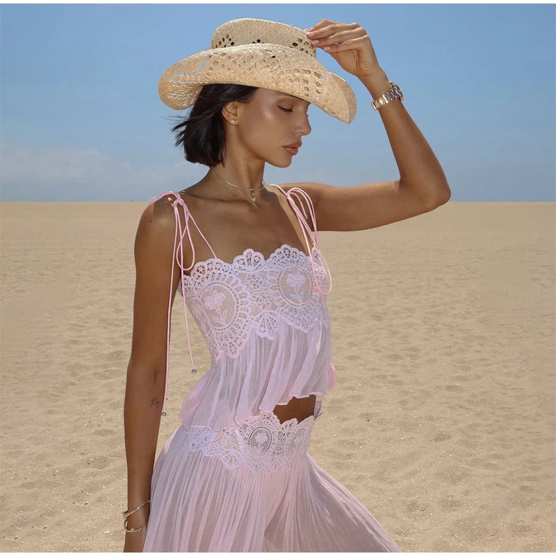 A woman standing on a sandy beach wearing a light pink sleeveless lace dress with thin straps and a wide-brim straw hat against a blue sky.
