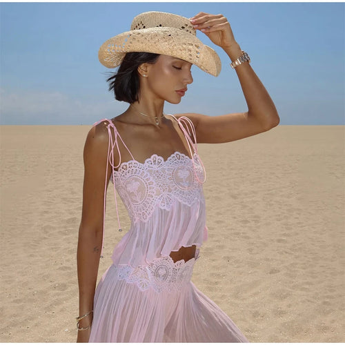 A woman standing on a sandy beach wearing a light pink sleeveless lace dress with thin straps and a wide-brim straw hat against a blue sky.