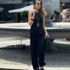A woman wearing a sleeveless black top with button details and wide-leg pants, both high-waisted. She is accessorized with sunglasses, a watch, and carrying a bag.
