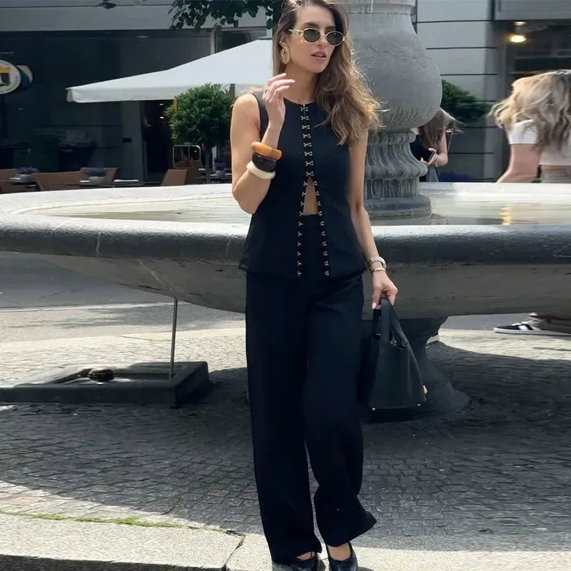 A woman wearing a sleeveless black top with button details and wide-leg pants, both high-waisted. She is accessorized with sunglasses, a watch, and carrying a bag.