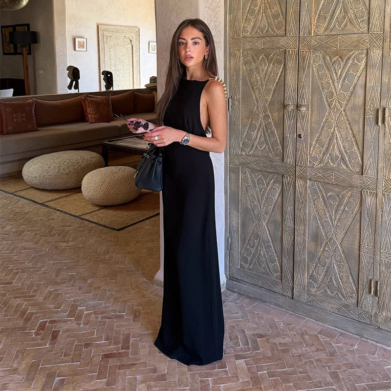 A woman stands indoors wearing a sleeveless black maxi dress with gold embellishments along the shoulder straps. She is holding a small black handbag and standing in a luxurious room with a large carved wooden cabinet, a terracotta tile floor, and a sofa in the background.