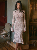 Woman wearing a lace dress standing in an elegant room with a mirror and chair.