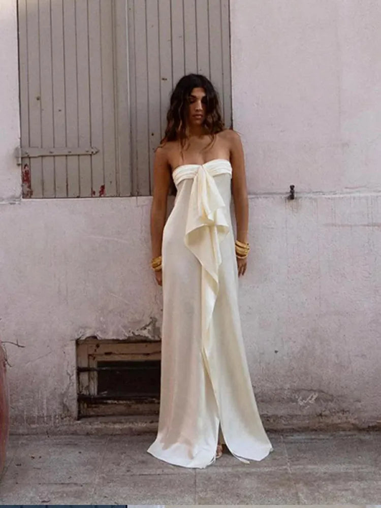 Woman wearing a strapless white gown with a bow detail against a plain wall.