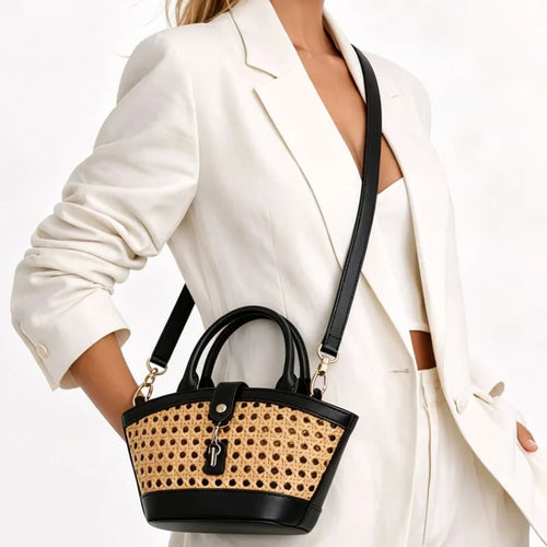A woman in a white blazer carries a small structured handbag with black leather trim and handles, plus a natural woven cane or raffia front panel and a black shoulder strap.