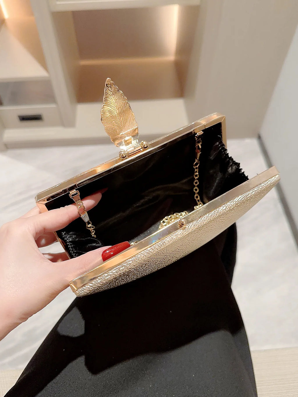 Gold clutch with feather design held by a hand against a neutral background