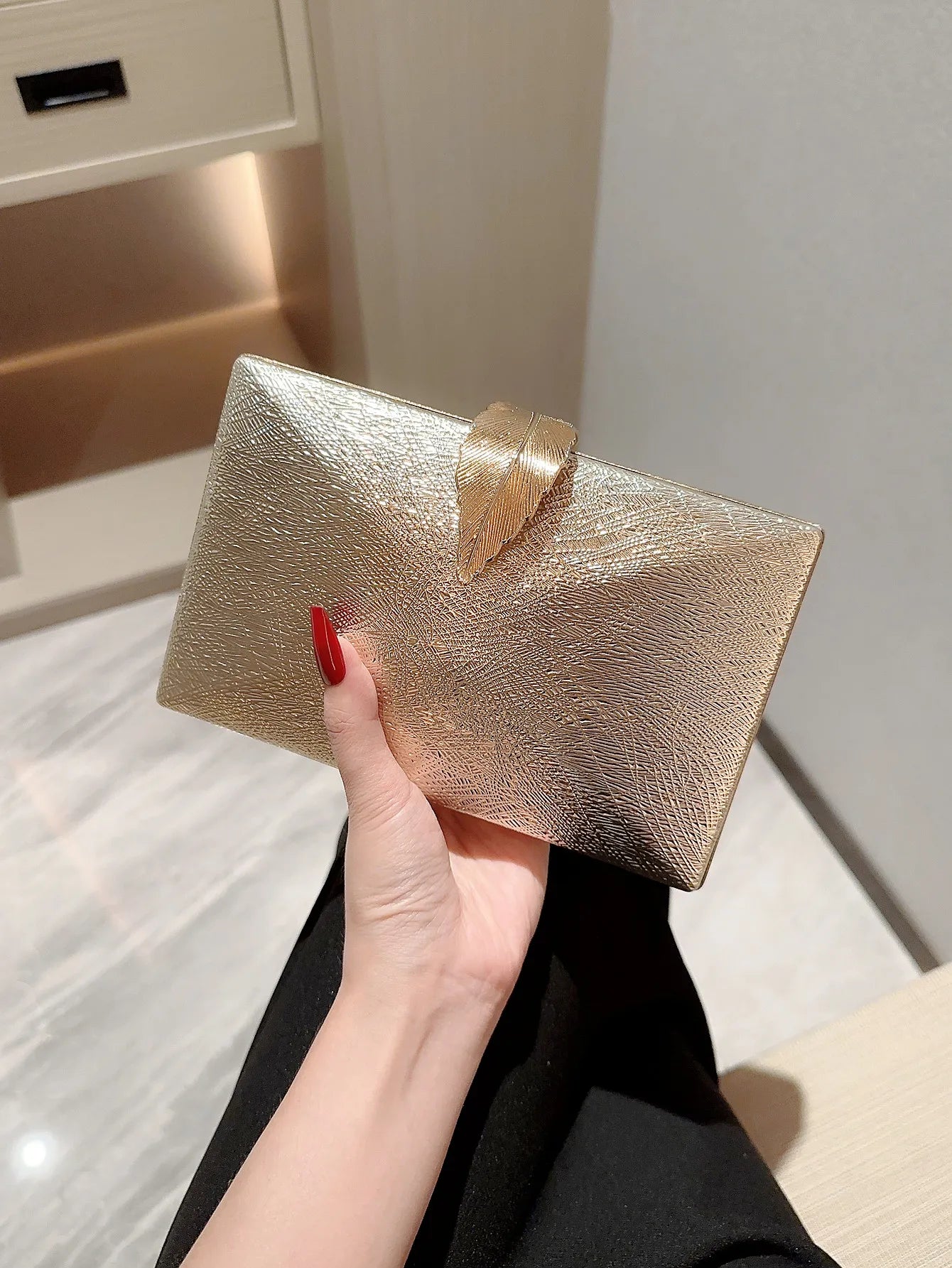 Gold clutch held by a hand with red nail polish against a neutral background