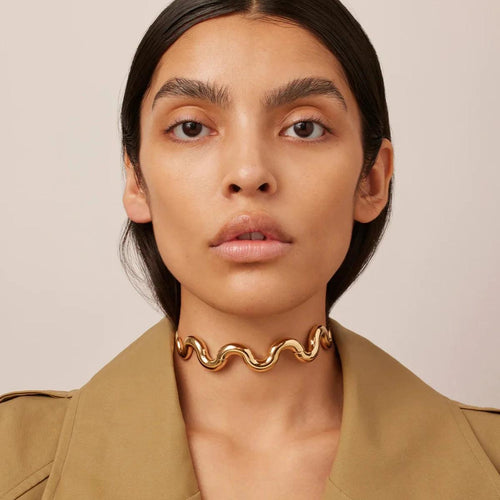 A person wearing a gold-colored wave-shaped choker necklace.
