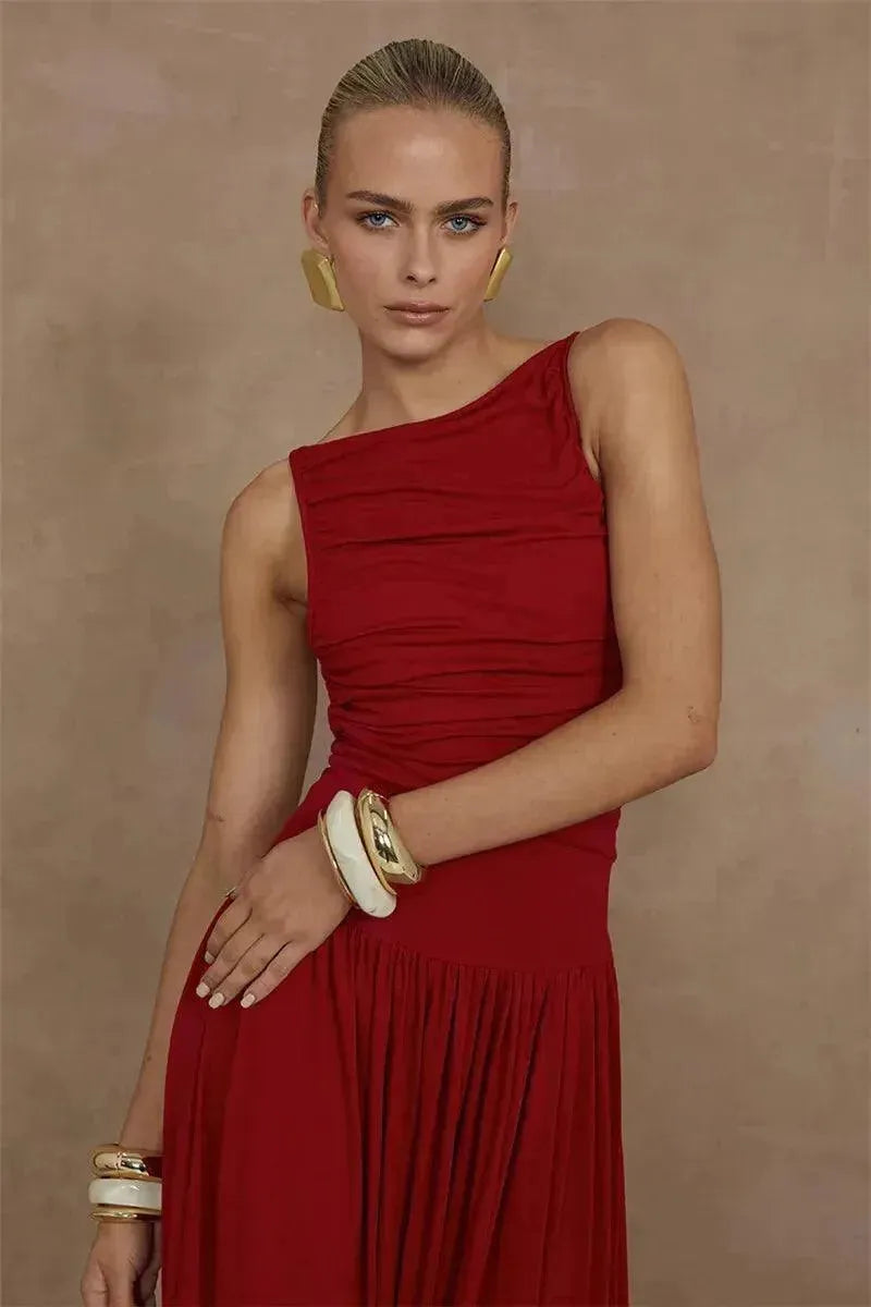 A woman wearing a red maxi dress with a boat neckline, reaching her thighs. She is also wearing a pair of gold-toned earrings and has a tan wristwatch.