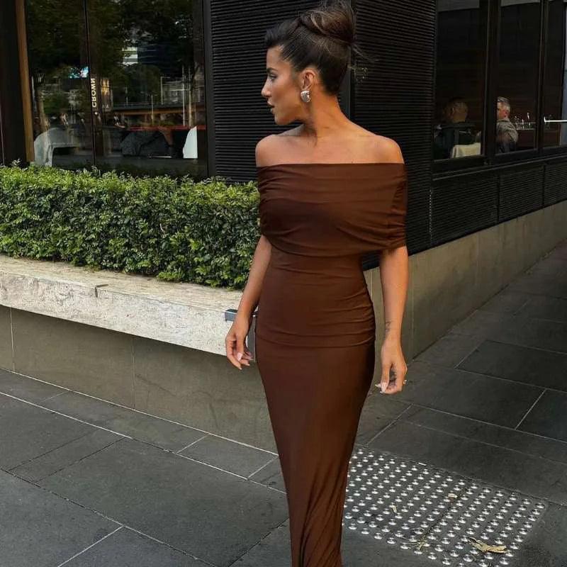 A woman wearing a brown off-the-shoulder maxi dress with a twist back design, standing on the sidewalk.