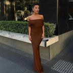 A woman wearing a brown off-the-shoulder maxi dress with a twist back design, standing on the sidewalk.