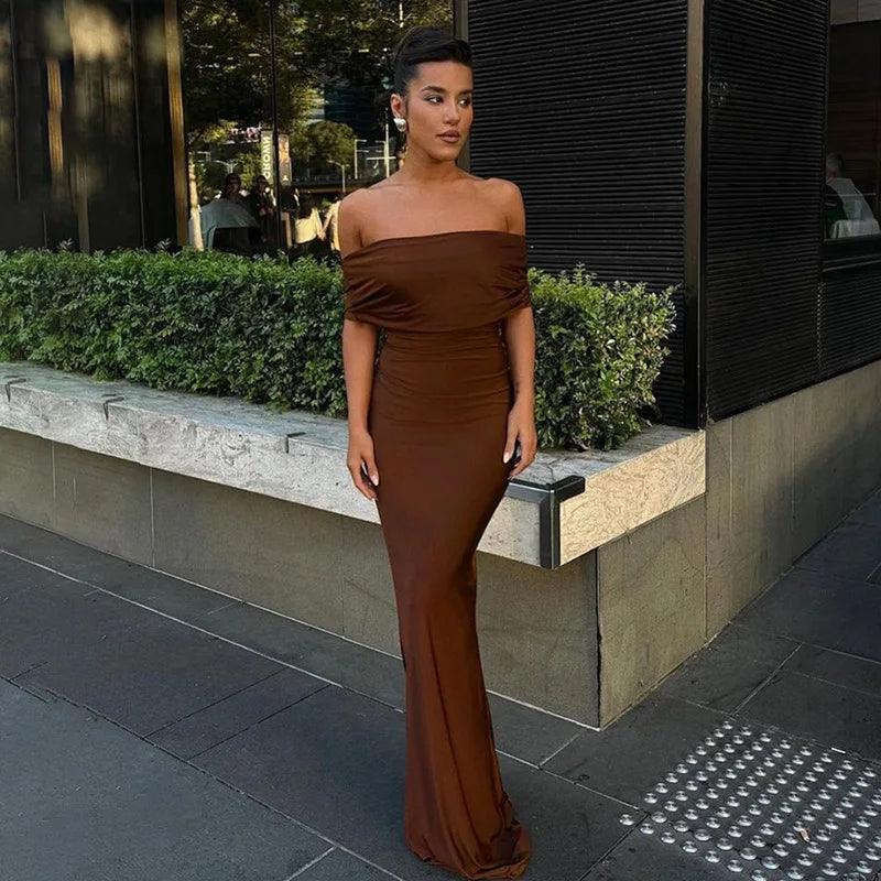 A woman wearing a brown off-the-shoulder maxi dress with a twist back design, standing on the sidewalk.