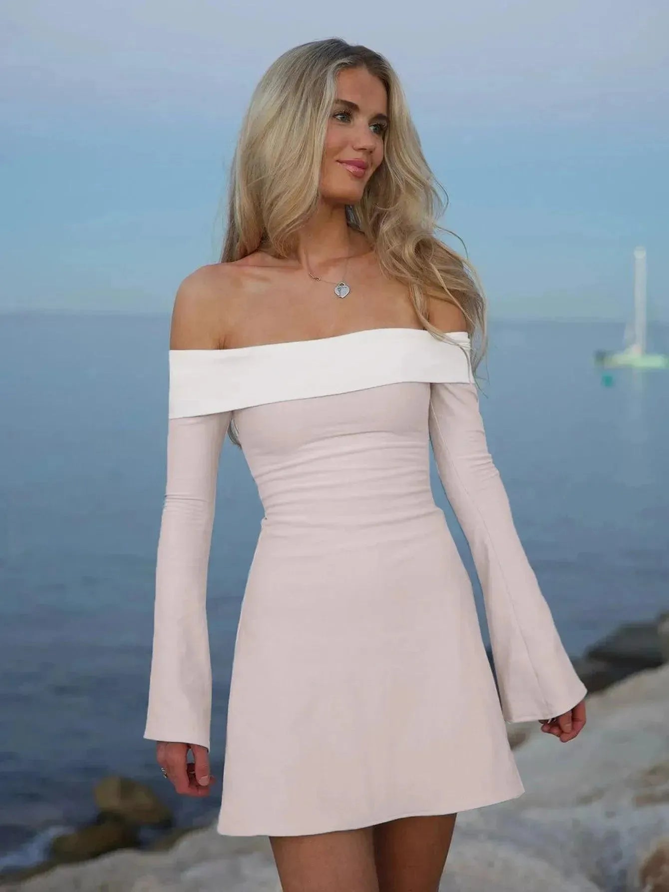 A woman wearing a pink, long sleeve, off-shoulder mini dress with a flare above the knee, standing by a body of water.
