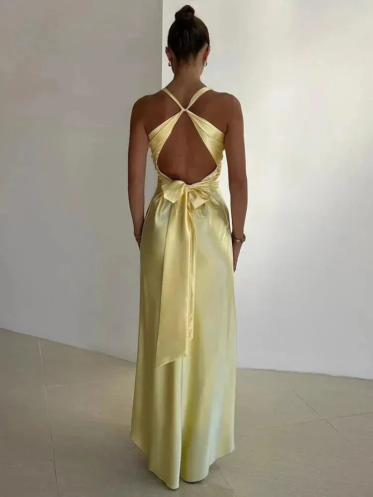 Woman in a light yellow evening gown with open back and ribbon details, standing against a plain background.