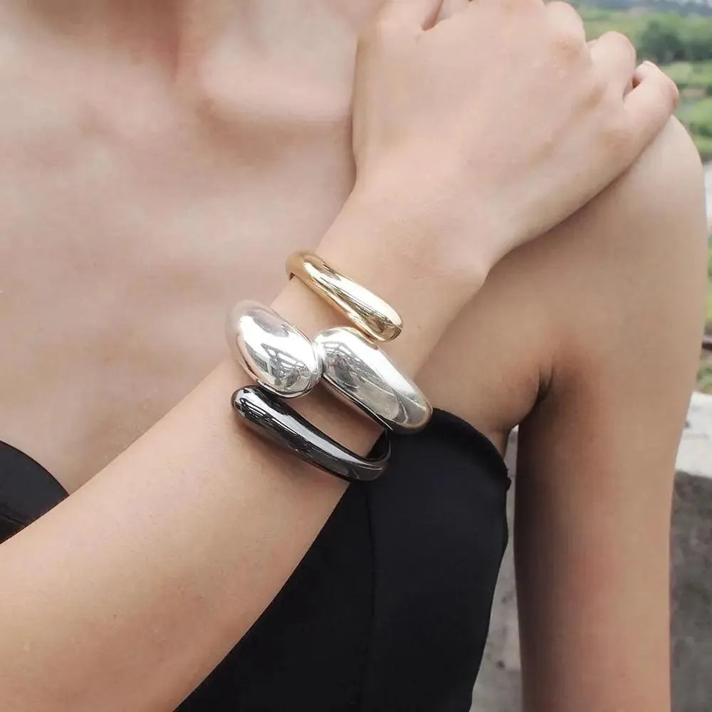 A person wearing two cuff bracelets, one with a gold-colored plating and the other with a silver-colored plating, on their left wrist.