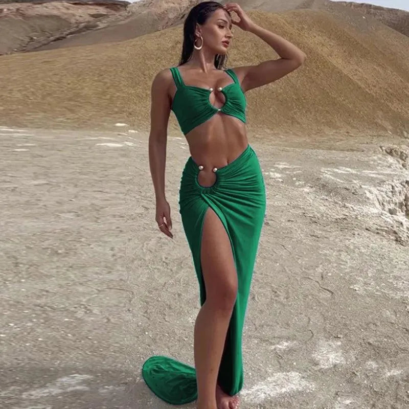 A female model wearing a green sleeveless top and a matching green maxi skirt with an asymmetrical design, standing in a sandy outdoor setting.