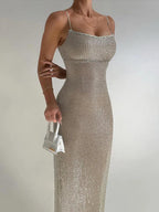 A model wearing a metallic silver, V-neck, spaghetti strap dress with a mid-calf length. The dress is adorned with sparkles, and the model is holding a small white bag.