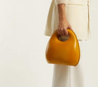 A woman holding a yellow, round flap handbag with a zipper closure.