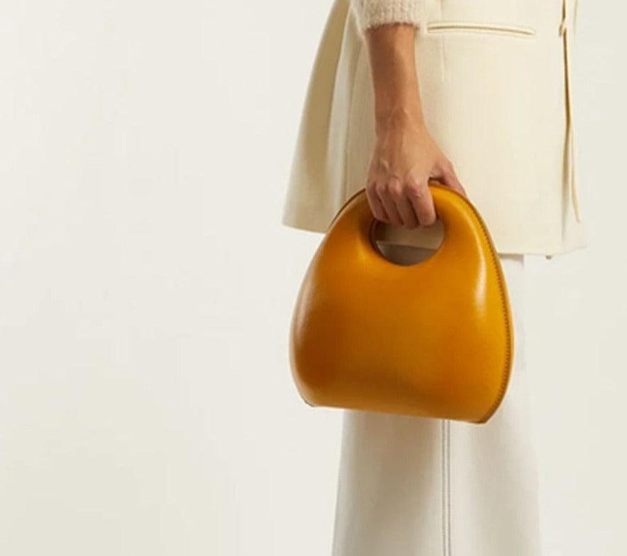 A woman holding a yellow, round flap handbag with a zipper closure.